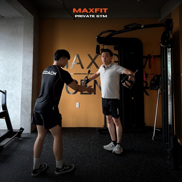 A high-contrast 1:1 image showing a personal trainer providing instruction to a client on a cable crossover machine.